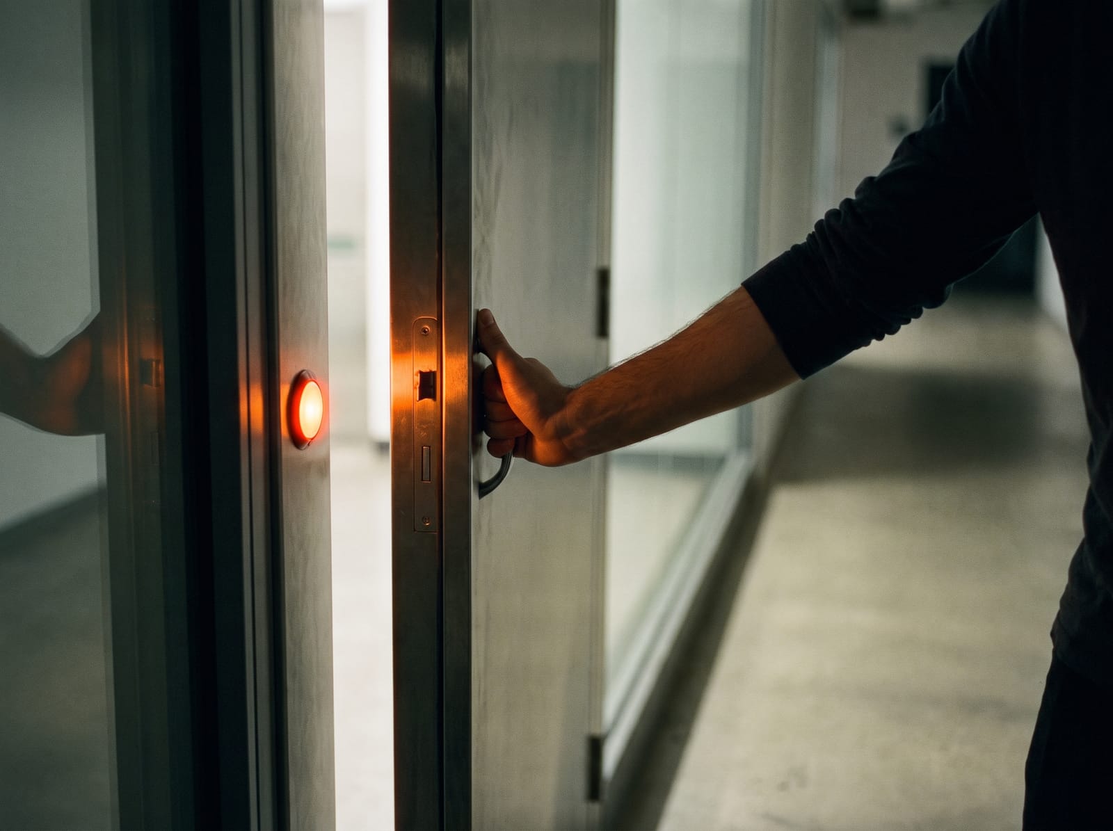 A hand holding open a steel service door into a sunlit office corridor